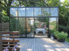 Contemporary 8x10x9 ft greenhouse with a sloping roof, placed on a wooden deck with a person dining amidst potted plants.