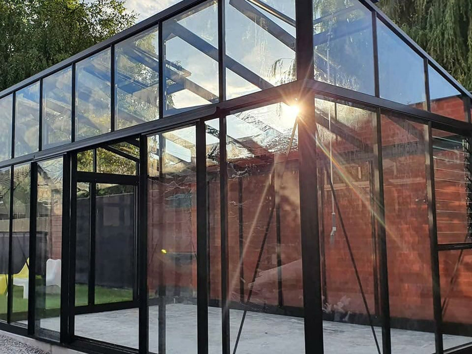 The Exaco Janssens modern greenhouse with a sloped roof captures the warm glow of evening lights, inviting a cozy atmosphere for an intimate garden dinner.
