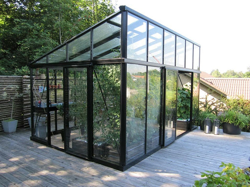 Exaco Janssens modern greenhouse with a sleek sloped roof, standing on a wooden deck surrounded by lush greenery, offering an ideal space for year-round gardening.