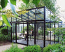 Modern M23 greenhouse with sloping roof and black frame, set in a lush garden with a person enjoying a quiet read inside.