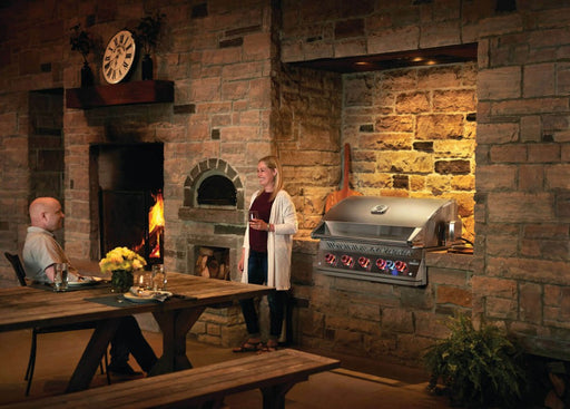 A cozy lifestyle setting featuring the Napoleon Grills Built-In 700 Series 38-Inch Gas Grill Head alongside a rustic stone fireplace, with people enjoying the warmth and a grilled meal.