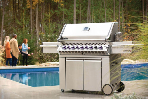 Napoleon Grills Prestige PRO™ 665 RSIB Grill in use with purple illuminated control knobs and a group of people in the background enjoying the outdoors.