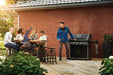 The Napoleon Grills Rogue XT 625 SIB Grill in an outdoor setting on a wooden deck with a group of people dining and one person operating the grill.