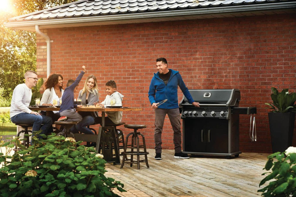The Napoleon Grills Rogue XT 625 SIB Grill in an outdoor setting on a wooden deck with a group of people dining and one person operating the grill.