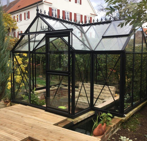 Janssens Exaco Orangerie Greenhouse with traditional black frame and clear panes, built on a wooden deck next to a pond in a residential backyard.