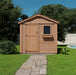 A wooden 8x8 Outdoor Living Today Gardener Shed with single door, situated on a grassy lawn beside a stone pathway, under a clear sky.