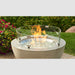 A 20-inch round glass wind guard in use, containing flames within a fire pit, set against a garden background.
