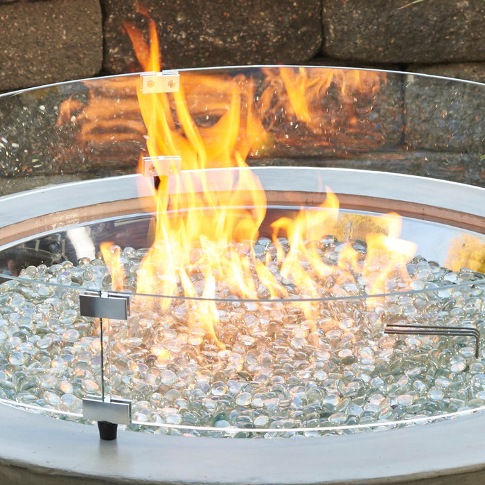 A round glass wind guard effectively containing flames on an outdoor fire pit with a garden backdrop.