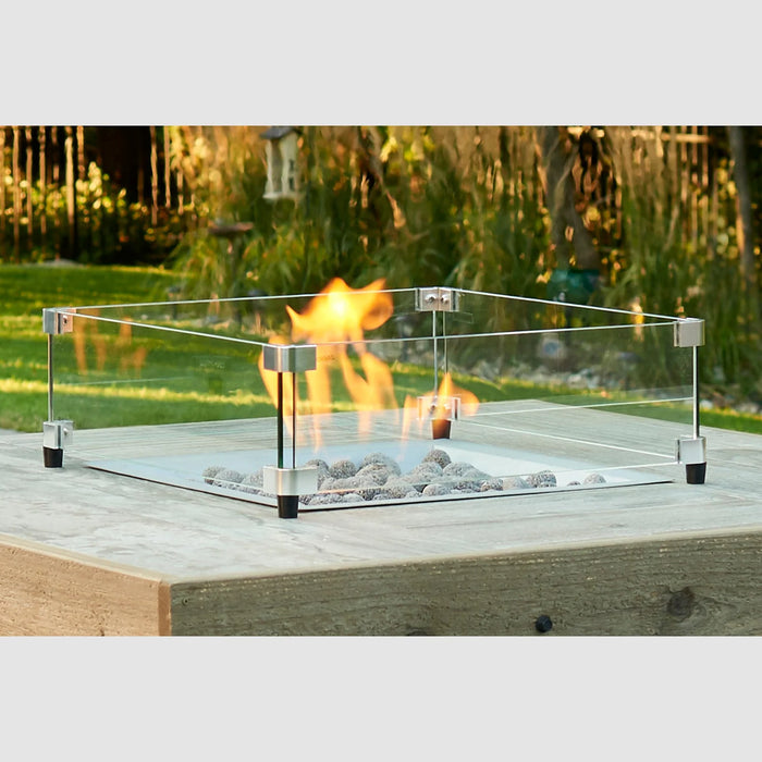 A 16x16 inch square glass wind guard containing flames on an outdoor fire pit with a lush garden in the background.