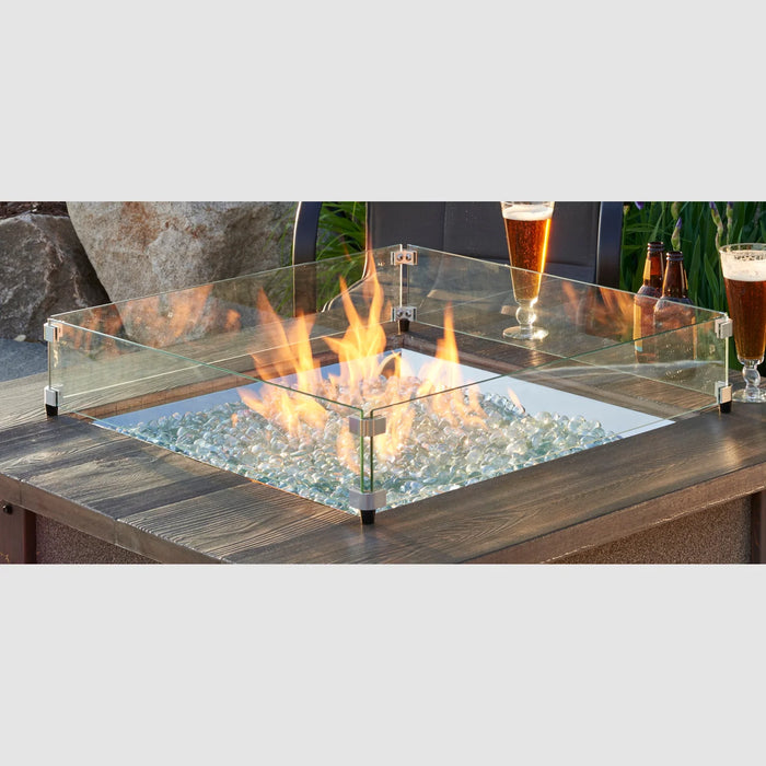 A 24x24 inch square glass wind guard in use over a fire pit, with flames visible and beverages on the deck in the foreground.