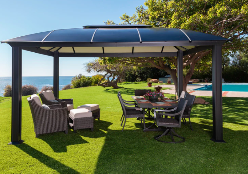 The Paragon Outdoor Cambridge Hard Top Gazebo 11x16 set in a lush seaside garden with a dining area underneath, offering a picturesque view of the sea horizon.