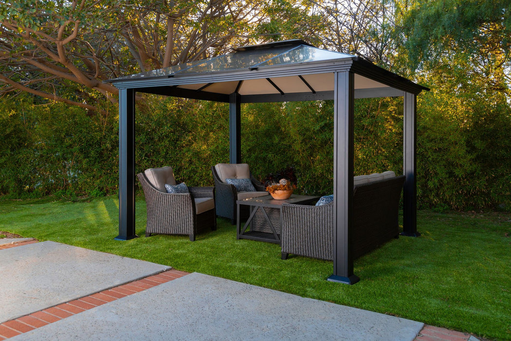 12x12 Santa Monica Hard Top Gazebo set in a lush garden, furnished with a cozy wicker love seat and chairs with plush cushions.