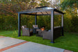 12x12 Santa Monica Hard Top Gazebo set in a lush garden, furnished with a cozy wicker love seat and chairs with plush cushions.