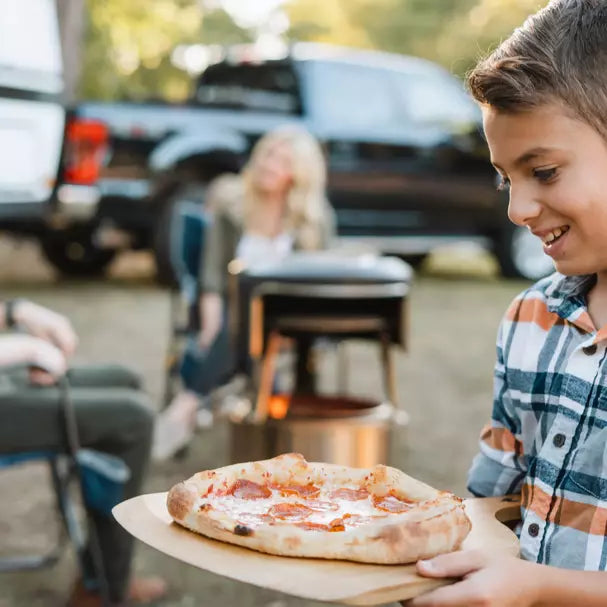 A boy holding pizza with Pi Fire Fire Pit Pizza Oven in the background.