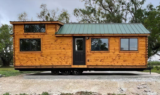 A brown, wooden Pinecrest Cabin with a green metal roof sits on a trailer. It has several windows and a black door.