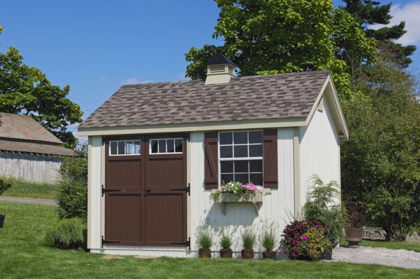 View of the Colonial Pinehurst shed kit by Little Cottage Company set in a garden, featuring cream siding with brown doors and window shutters, complemented by a flower box.