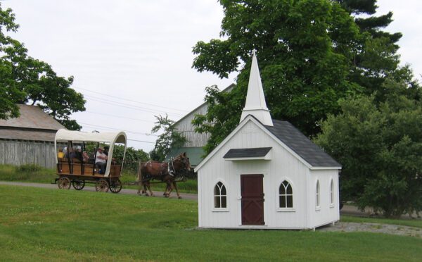 The Little Cottage Company's Chapel Playhouse in a meadow, as a traditional horse-drawn carriage passes by on a sunny day.