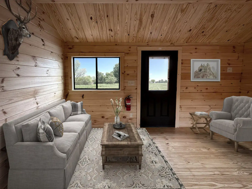 A cozy room in the Oasis Tiny Estates Ranger Cabin features a gray couch, armchair, coffee table, wall-mounted deer head, rug, fire extinguisher, a window with an outdoor view, and a framed picture on the wall.