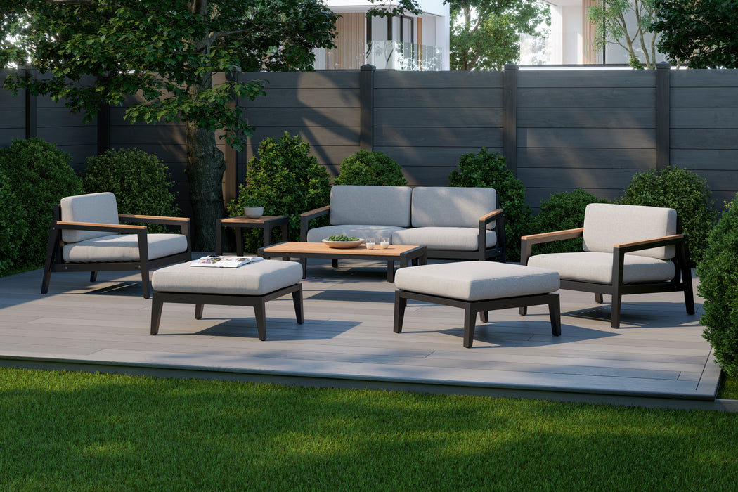 Rhodes 6 Seater Chat Set arranged on a patio with coffee table and side table, featuring cushioned seats and wooden frames against a backdrop of lush greenery and a grey fence.