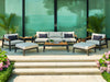 Rhodes Furniture Set arranged on a patio, featuring two armchairs, a sofa, two ottomans with cushions, and a center table, framed by lush greenery.