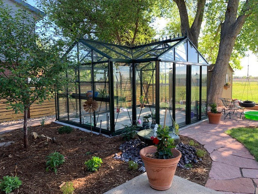 Tranquil backyard installation of an Exaco Janssens Royal Victorian Orangerie Greenhouse with clear glass panes and black framing, nestled among trees and potted plants.