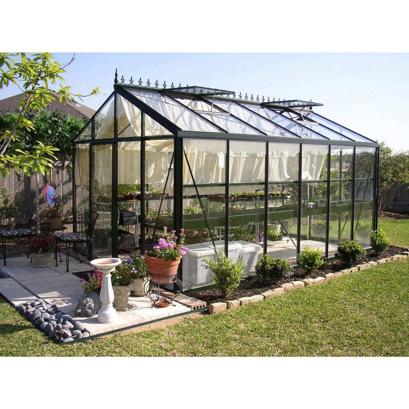 A Royal Victorian VI-34 greenhouse on a patio with open curtains and surrounding foliage in a serene garden setting.