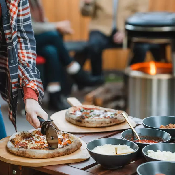 A person cutting pizza with Pi Fire Fire Pit Pizza Oven in the background.