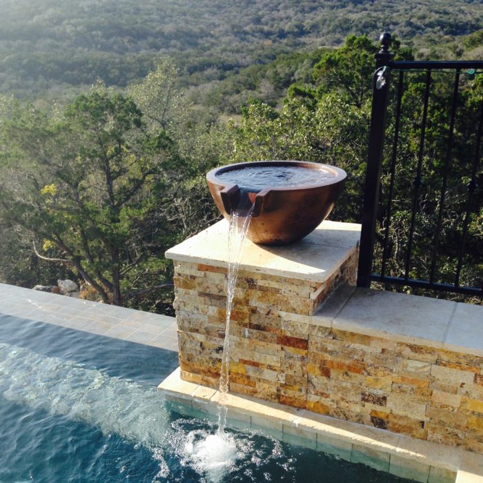 A Slick Rock Concrete Spill Water Bowl in a weathered copper finish acts as a water feature, with water cascading from its spout into a pool below. The bowl is positioned on a stone pedestal beside a swimming pool with a scenic hill view in the background.