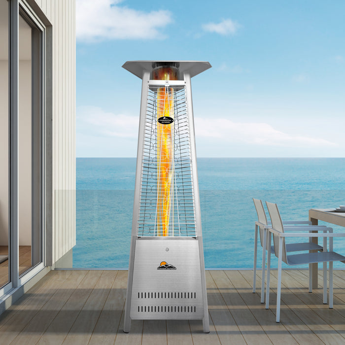 The image shows a stainless steel Inferno Flame Tower Heater situated on a wooden deck by the beachside.