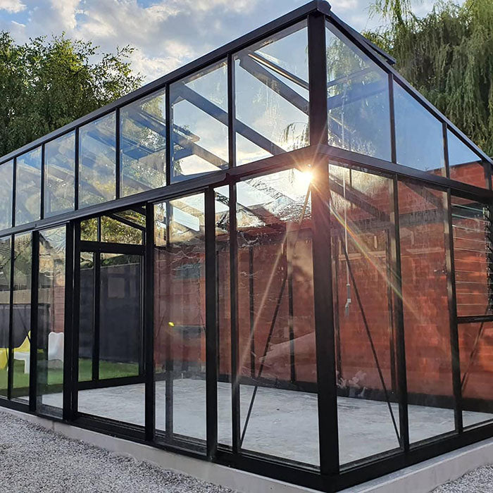 A striking Exaco Janssens Modern M23 Greenhouse captures the sunset's rays through its transparent glass and sloping roof, accentuating the sturdy black frame against a serene backyard setting.