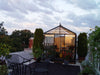 An Exaco Janssens Royal Victorian VI 23 Greenhouse stands on a rooftop patio, adorned with plants and lights amidst outdoor furniture, set beneath a cloudy evening sky.