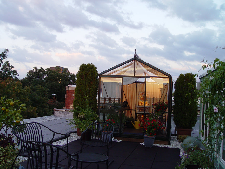 An Exaco Janssens Royal Victorian VI 23 Greenhouse stands on a rooftop patio, adorned with plants and lights amidst outdoor furniture, set beneath a cloudy evening sky.