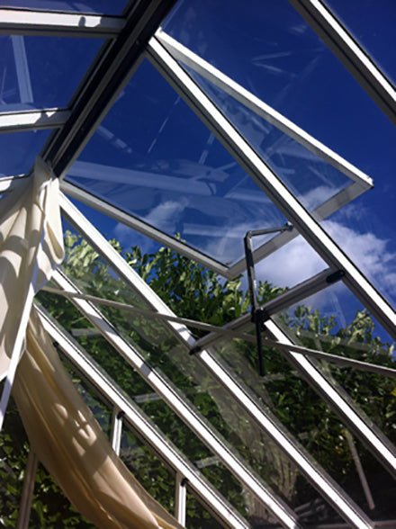 A view looking up at the roof window of the Antique Janssens greenhouse, with an open ventilation pane set against a blue sky.