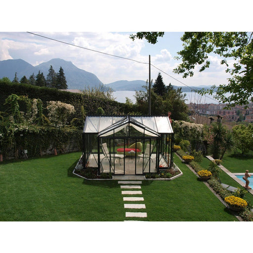 Lush garden view with Exaco Janssens Royal Victorian Orangerie Greenhouse featuring a transparent sloped roof and elegant frame against a scenic mountain backdrop.