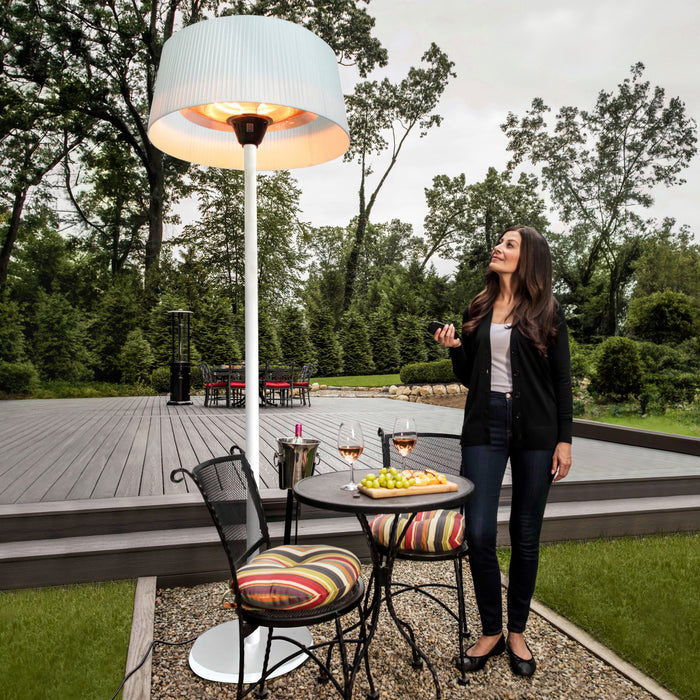 A woman stands on an elegant wooden deck next to a table with snacks and wine glasses, enjoying the warmth from the white Sol Electric Standing Heater. 