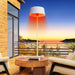 The Standing heater is on a balcony against a backdrop of a stunning sunset over a coastal landscape, highlighting its ambient lighting feature.