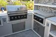 Wildfire outdoor kitchen setup featuring a stainless steel grill and griddle, sleek cabinetry, and marble-style countertops, set against a textured backsplash in a backyard with lush greenery.