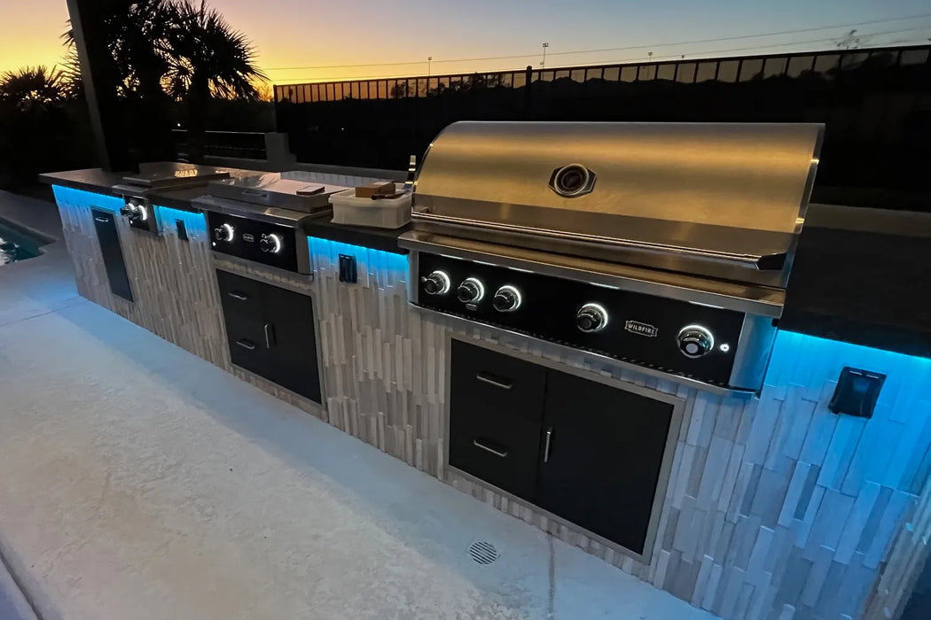 Wildfire outdoor kitchen setup featuring a grill, griddle, and sleek cabinetry with ambient blue lighting, set against a sunset backdrop by the poolside.