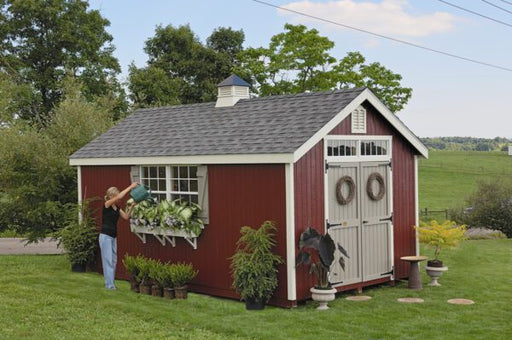 Person watering flowers on the side of the Colonial Williamsburg shed by Little Cottage Company with a red exterior and country-style accents.