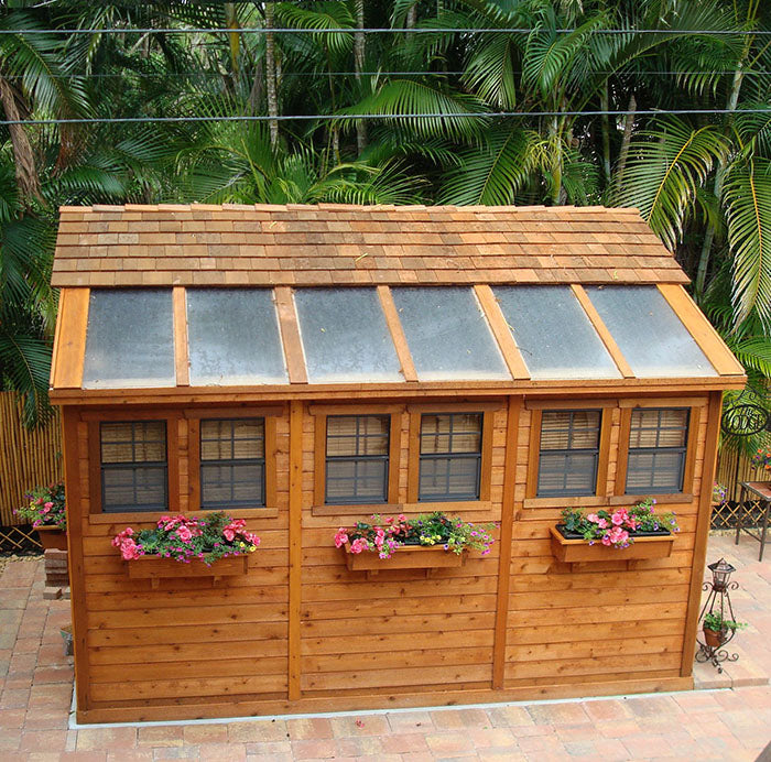 Outdoor Living Today Sunshed Garden Shed, 8x12, with cedar shingles and skylights, adorned with flower boxes, against a lush tropical background