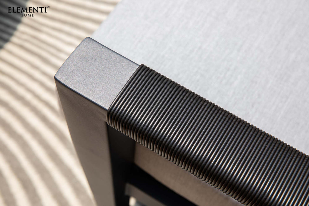 Close-up of Elementi's Laguna Collection chair—weather-resistant black armrest and light fabric seat highlight this stylish outdoor furniture. Shadows fall on a striped surface below, featuring the "Elementi" logo.