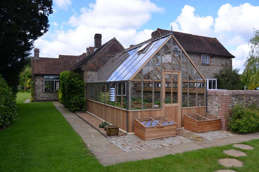 A Westminster cedar greenhouse situated in a garden beside a brick house, surrounded by green plants and two wooden cold frames in front.