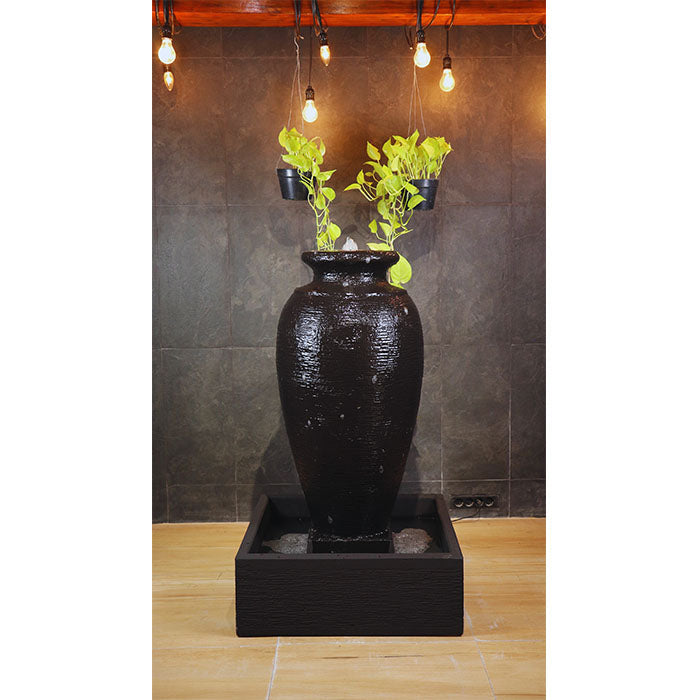 Amphora garden water feature with square basin and floating succulents, placed on green lawn with blooming flowers.
