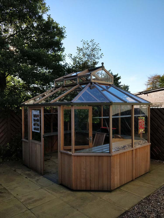 The Alton Cedar Structure Evolution Octagonal a wooden greenhouse with glass panels, is situated on a paved area in the garden. A "For Sale" sign is displayed on one side, with trees and a fence visible in the background under a clear blue sky.