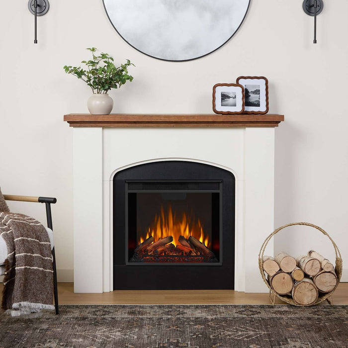 Real Flame Anika electric fireplace with black firebox surround in neutral living room, wood mantel with plant and framed photos, log basket, round mirror, and wall sconces above