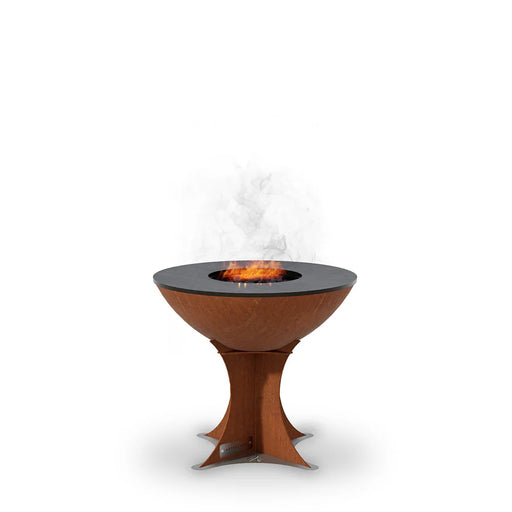 A round Arteflame grill with a tall Euro base stand, featuring a wood fire burning on its black cooking surface, emitting smoke.