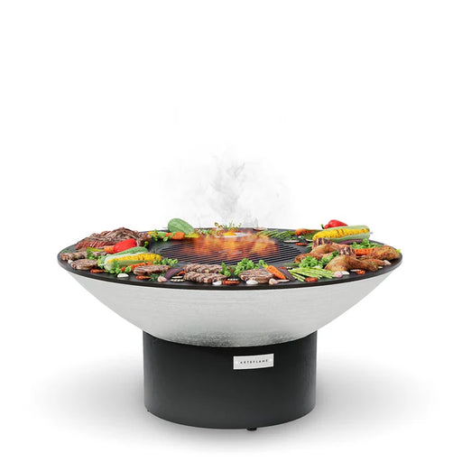 The Arteflame 40" Platinum Series Fire Pit With Cooktop host a modern, round grill brimming with a variety of meats and vegetables sizzling on its surface. Wisps of smoke rise gracefully from the hot food.