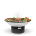 The Arteflame 40" Platinum Series Fire Pit With Cooktop host a modern, round grill brimming with a variety of meats and vegetables sizzling on its surface. Wisps of smoke rise gracefully from the hot food.