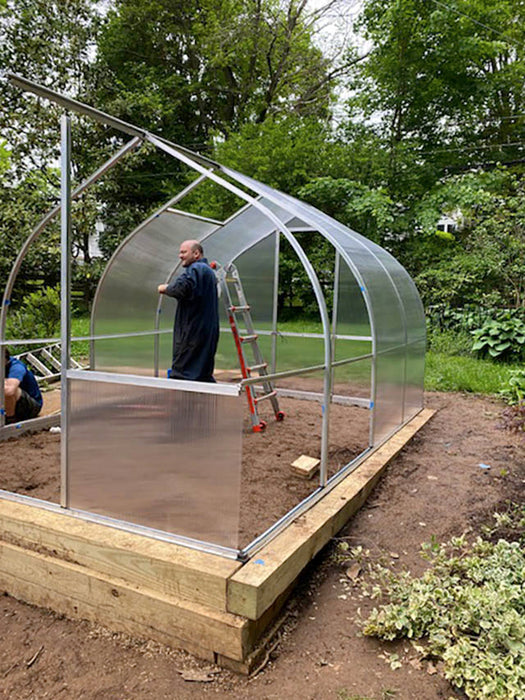 Homeowners assembling the aluminum frame of an Exaco Riga greenhouse, showing polycarbonate side panels and ladders during installation process.