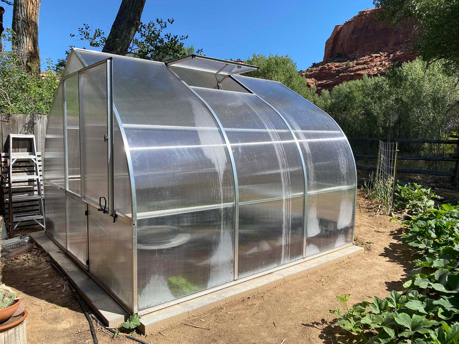 Close-up angle of Exaco Riga 3 greenhouse front door, showing durable aluminum frame, polycarbonate walls, and roof vent with garden in background.